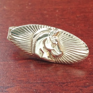 Puede incluir: Un broche de corbata dorado con un diseño de cabeza de caballo. La cabeza del caballo está de perfil y tiene una crin detallada. El fondo del broche de corbata es un patrón de rayos de sol.