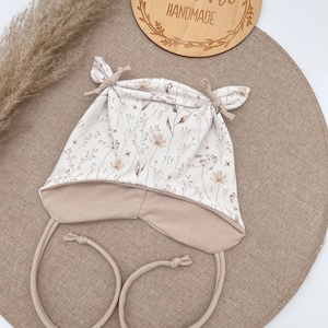 Op de afbeelding: Een beige baby muts met een bloemenprint en een strik aan de bovenkant. De muts heeft een beige voering en een lange strik die onder de kin kan worden geknoopt. De muts is handgemaakt en heeft de tekst "Emilima Handmade" op een houten bord op de achtergrond.