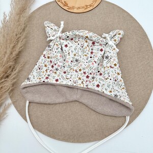 Könnte beinhalten: Ein Baby-Hut mit floralem Muster in Weiß, Burgunderrot und Gelb. Der Hut hat ein beiges Fleecefutter und weiße Bindebänder. Das Holzschild über dem Hut trägt die Aufschrift "Emilima Handmade".