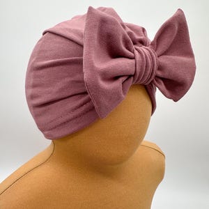 Könnte beinhalten: Ein mauvefarbener Turban mit einer großen Schleife oben. Der Turban ist aus einem weichen, dehnbaren Stoff.