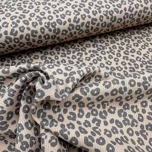 May include: Tan and gray leopard print fabric. The fabric has a ribbed texture and is folded in a layered pattern. The leopard spots are a dark gray color and are scattered across the tan background. The fabric is likely used for clothing or home decor.