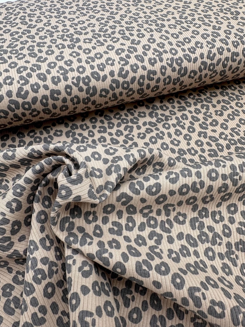 May include: Tan and gray leopard print fabric. The fabric has a ribbed texture and is folded in a layered pattern. The leopard spots are a dark gray color and are scattered across the tan background. The fabric is likely used for clothing or home decor.