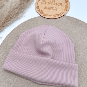 Könnte beinhalten: Eine hellviolette Rippstrick-Beanie-Mütze mit umgeschlagenem Saum. Die Mütze ist aus einem weichen, dehnbaren Material gefertigt. Ein Holzschild mit dem Text "Emilima Handmade" befindet sich im Hintergrund.