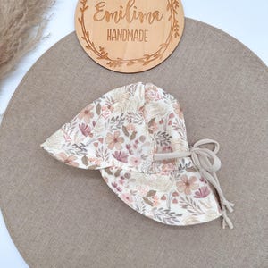 Könnte beinhalten: Ein Sonnenhut mit cremefarbener Basis und einem Blumenmuster in Rosa-, Braun- und gedeckten Grüntönen. Der Hut hat eine breite Krempe und seitliche Stoffbänder in Beige zum Binden. Der Stoff ist mit einem botanischen Druck versehen.