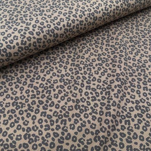 May include: Tan fabric with a dark gray leopard print pattern. The fabric has a ribbed texture, and the leopard spots are irregularly shaped and scattered across the surface. The fabric is rolled up, showing the pattern in detail.