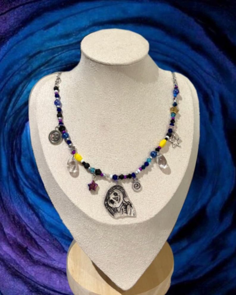 May include: A beaded necklace with a variety of charms, including a star, a clear crystal, and a circular pendant. The beads are in shades of blue, purple, yellow, and black. A small, illustrated pendant of a character is also included.