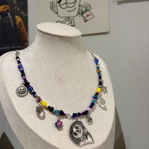 May include: A beaded necklace with a variety of charms, including a button, a star, and a clear teardrop. The necklace features blue, yellow, and purple beads. A pendant with a cartoon face hangs from the necklace.