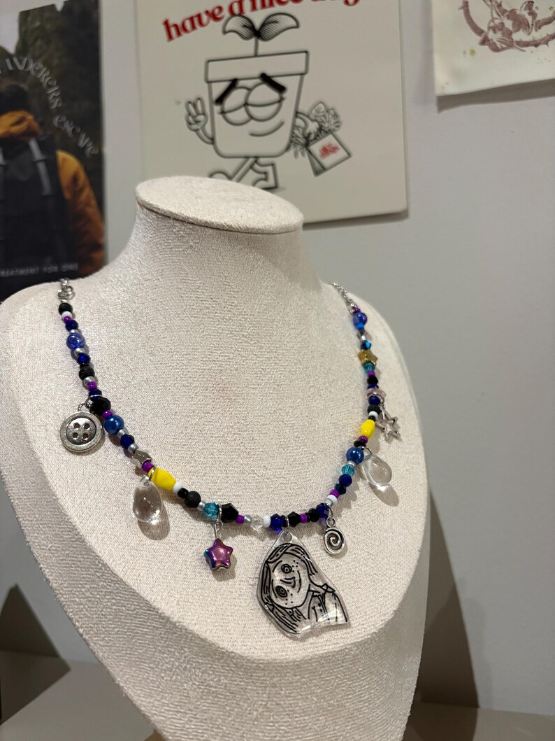 May include: A beaded necklace with a variety of charms, including a button, a star, and a clear teardrop. The necklace features blue, yellow, and purple beads. A pendant with a cartoon face hangs from the necklace.