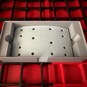 Puede incluir: Un objeto rectangular gris claro con múltiples agujeros pequeños, dentro de una bandeja gris claro. La bandeja está sobre una superficie roja y negra en forma de rejilla. El objeto parece ser una almohadilla de lijado.