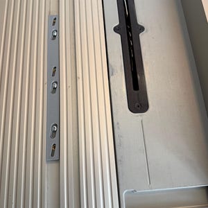 May include: Close-up of a silver table saw with a black blade guard and a gray metal fence. The saw has a ribbed surface and a ruler marked in inches.