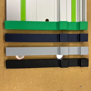 May include: A close-up shot of four different colored guide rails. The top rail is green, followed by navy blue, gray, and black. The rails are rectangular with a square block on each end. The background is a light brown surface.