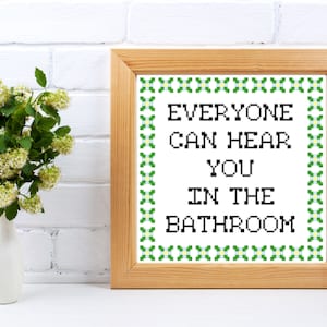 Peut inclure: Un cadre en bois clair contenant une broderie au point de croix avec le texte "EVERYONE CAN HEAR YOU IN THE BATHROOM" en noir. Le cadre est orné d'une bordure verte. Un vase blanc avec des fleurs est à gauche.