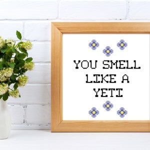 May include: A framed cross-stitch art piece with the text "YOU SMELL LIKE A YETI" in black pixelated letters. The frame is light brown. The artwork is decorated with small blue flowers. A white vase with green and white flowers is to the left.