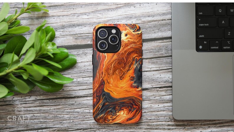 Puede incluir: Una funda de tel&eacute;fono con un dise&ntilde;o abstracto vibrante en tonos naranja, marr&oacute;n y negro. La funda tiene un acabado brillante y un recorte para la c&aacute;mara. La funda del tel&eacute;fono est&aacute; sobre una superficie de madera con hojas verdes y un port&aacute;til.