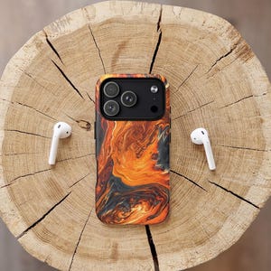 Puede incluir: Una funda de tel&eacute;fono con un dise&ntilde;o naranja y negro llameante, y auriculares inal&aacute;mbricos blancos se muestran sobre una superficie de madera. La funda del tel&eacute;fono tiene un m&oacute;dulo de c&aacute;mara con tres lentes. El logotipo de Craft House Cases es visible.