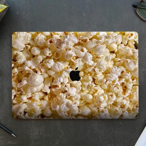 May include: A laptop computer with a popcorn design skin. The skin is a close-up of popcorn kernels, and the Apple logo is visible on the laptop.