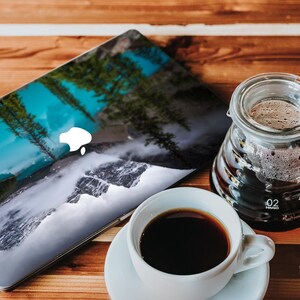 May include: A laptop computer with a scenic nature image on the lid. The image features a waterfall, mountains, and trees. A glass coffee pot and a white coffee cup are on a wooden table.