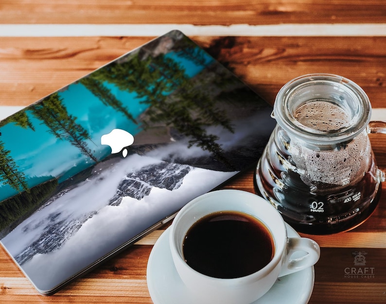 May include: A laptop computer with a scenic nature image on the lid. The image features a waterfall, mountains, and trees. A glass coffee pot and a white coffee cup are on a wooden table.