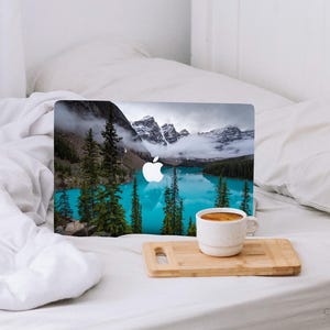 May include: A laptop computer with a scenic mountain lake image on the cover. The laptop is sitting on a white bed with a cup of coffee on a wooden cutting board.