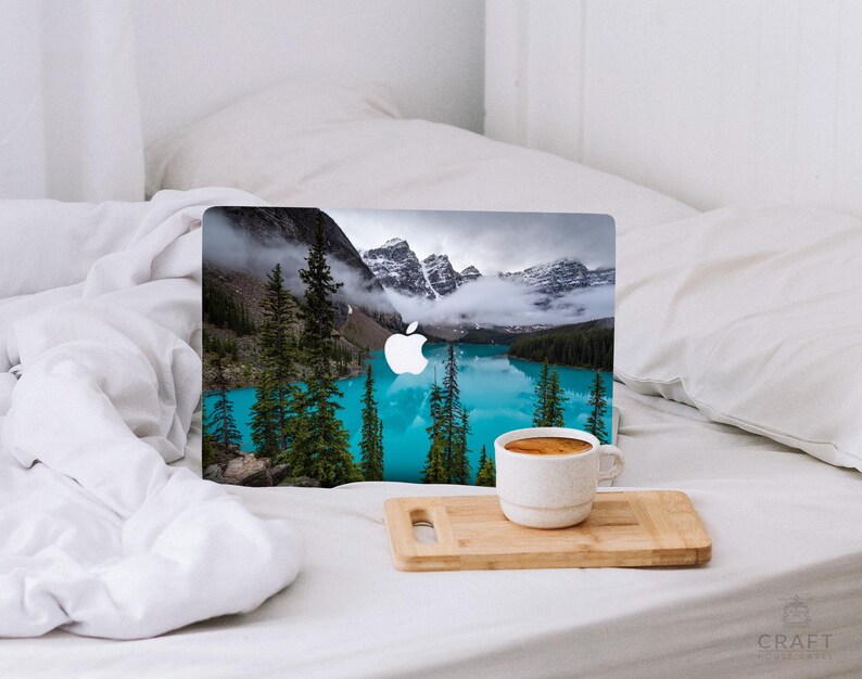 May include: A laptop computer with a scenic mountain lake image on the cover. The laptop is sitting on a white bed with a cup of coffee on a wooden cutting board.