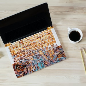 May include: A laptop computer with a colorful abstract design covering the keyboard and trackpad. The laptop is on a wooden surface with a cup of coffee and pencils.