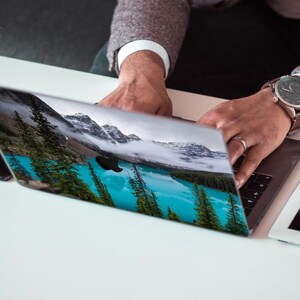 May include: A person is using a laptop with a scenic landscape image on the screen. The image shows a lake surrounded by trees and mountains. The laptop is on a white surface.