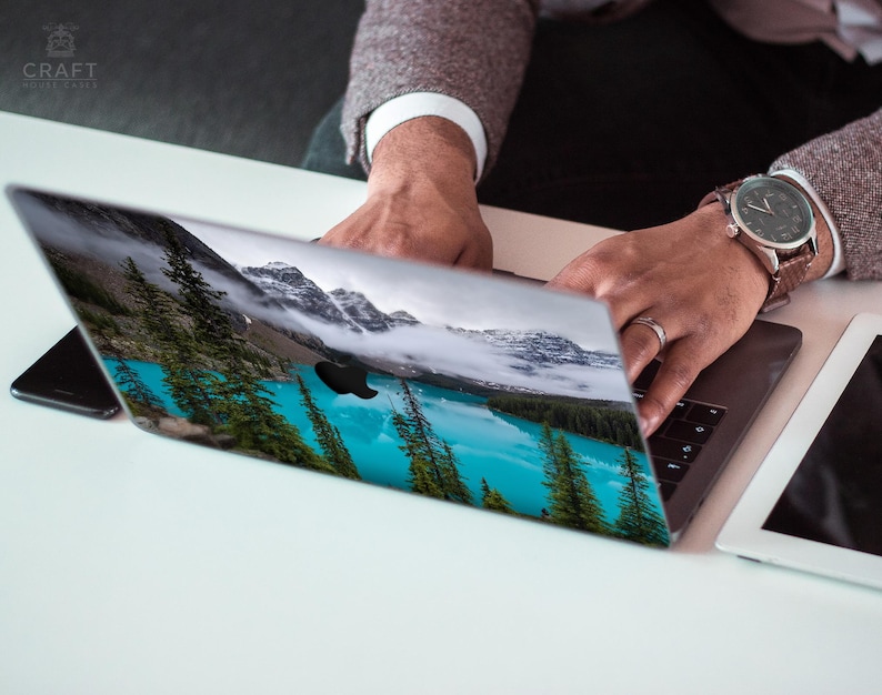 May include: A person is using a laptop with a scenic landscape image on the screen. The image shows a lake surrounded by trees and mountains. The laptop is on a white surface.
