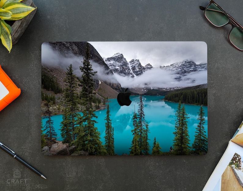 May include: A laptop skin with a scenic image of a mountain lake with snow-capped peaks and a blue sky. The lake is surrounded by trees and the water is a vibrant blue.