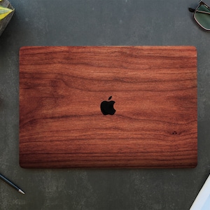 Può includere: Un computer portatile con una pelle con un motivo a grana di legno. La pelle è di colore marrone scuro e presenta un sottile motivo a grana di legno. Il logo Apple è visibile sul coperchio del laptop.