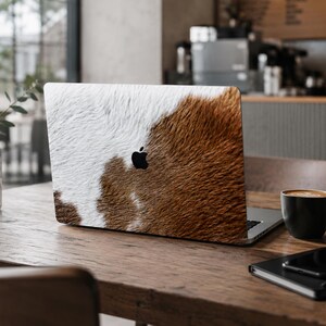 以下が含まれることがあります： シルバーのノートパソコンの蓋に貼られた、リアルな茶色と白の牛革の毛皮模様のスキン。デザインはデバイスの背面全体を覆っており、質感のあるアニマルプリントと中央の黒いAppleロゴが特徴です。