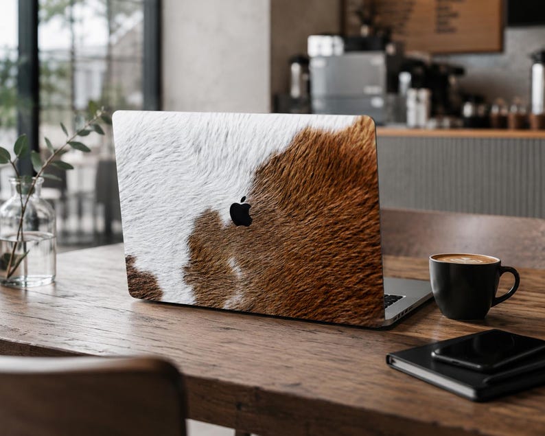 以下が含まれることがあります： シルバーのノートパソコンの蓋に貼られた、リアルな茶色と白の牛革の毛皮模様のスキン。デザインはデバイスの背面全体を覆っており、質感のあるアニマルプリントと中央の黒いAppleロゴが特徴です。