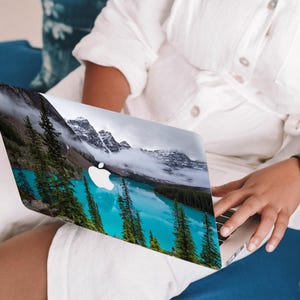May include: A laptop computer with a scenic mountain lake and forest landscape on the cover. The laptop is open and the screen is visible. The person's hand is resting on the keyboard.