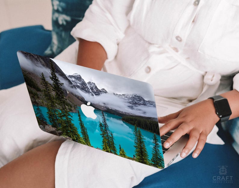 May include: A laptop computer with a scenic mountain lake and forest landscape on the cover. The laptop is open and the screen is visible. The person's hand is resting on the keyboard.
