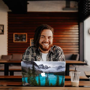 May include: A man with long brown hair and a beard smiles at the camera while sitting at a table with a laptop. The laptop has a sticker on it that shows a mountain lake with trees and a blue sky.