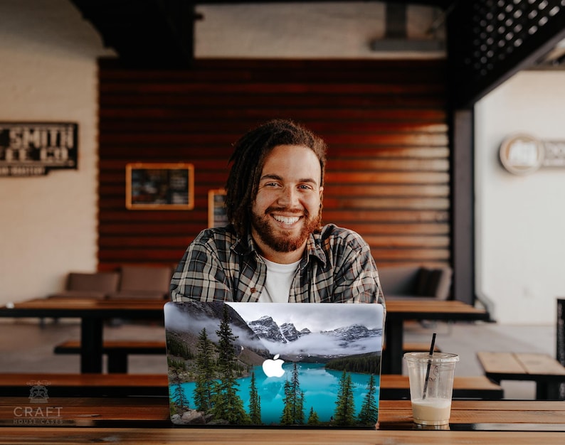 May include: A man with long brown hair and a beard smiles at the camera while sitting at a table with a laptop. The laptop has a sticker on it that shows a mountain lake with trees and a blue sky.