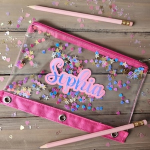 May include: A clear pencil pouch with a pink zipper and a pink reinforced strip featuring three metal grommets. The pouch contains loose heart and star-shaped confetti and displays the name Sophia in pink script. Two light pink pencils are included.