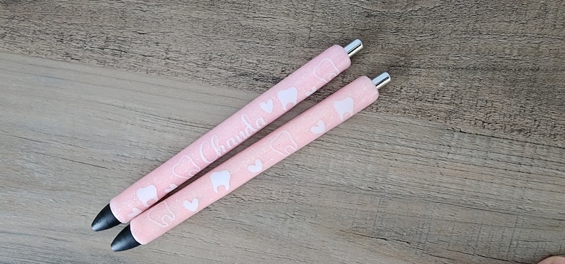May include: Two pink pens with white tooth and heart designs. The pen on the top has the name "Chanda" written on it.