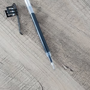 May include: A black and clear pen with a metal clip lying on a light brown wooden surface.