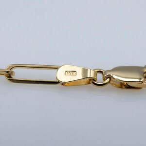May include: Close-up of a gold chain with a lobster clasp. The chain is made of small, rectangular links. The clasp is stamped 14K.