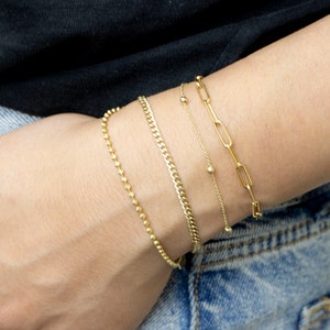 May include: Four gold bracelets stacked on a person's wrist. The bracelets are different styles, including a chain link, a beaded chain, and a curb chain.