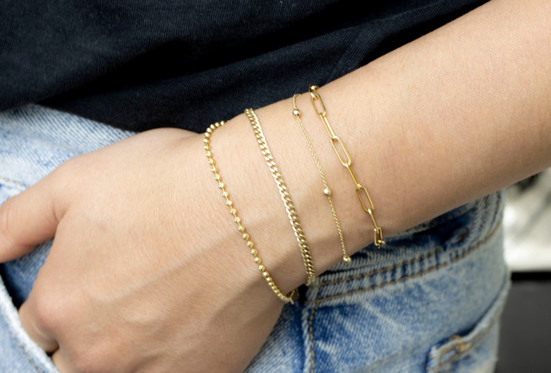 May include: Four gold bracelets stacked on a person's wrist. The bracelets are different styles, including a chain link, a beaded chain, and a curb chain.