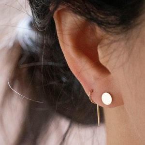 May include: A gold-toned earring set consisting of a small, flat, circular stud and a delicate chain that connects to a thin, straight, vertical bar component. The jewelry features a polished metallic finish and is worn in the earlobe.