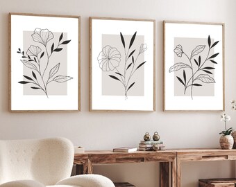 Puede incluir: Tres láminas botánicas enmarcadas. Cada lámina presenta un dibujo lineal negro de una flor o planta sobre un fondo beige claro, dentro de una forma rectangular. Los marcos son de color marrón claro. Las láminas se exhiben sobre una consola de madera.