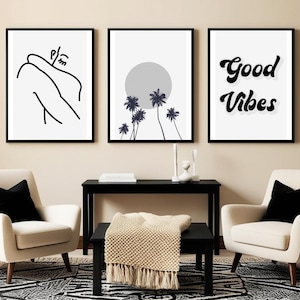 May include: Three framed art prints. The first features a black and white line drawing of a person. The second shows palm trees and a gray sun. The third says "Good Vibes" in black script. The prints hang above two cream armchairs with black pillows.