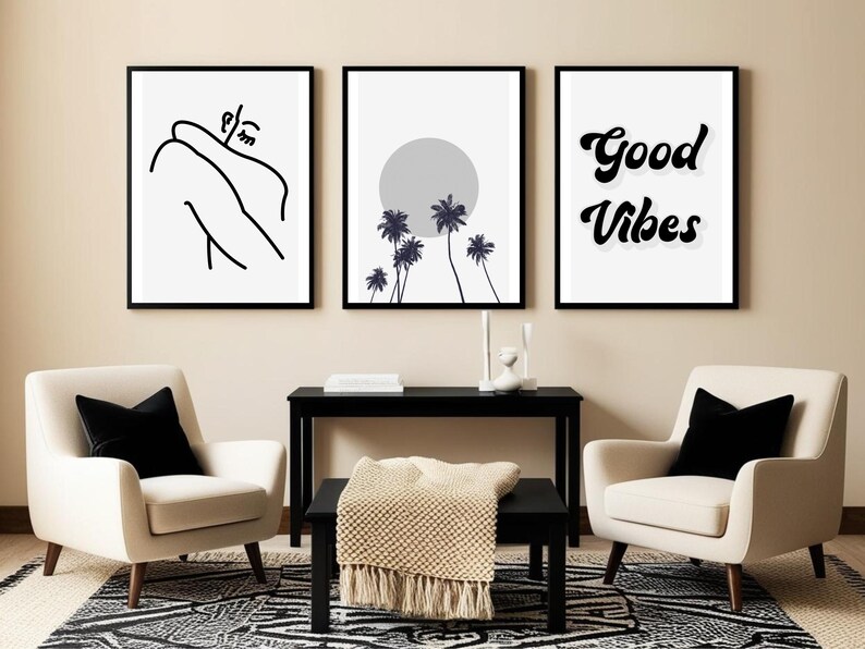 May include: Three framed art prints. The first features a black and white line drawing of a person. The second shows palm trees and a gray sun. The third says "Good Vibes" in black script. The prints hang above two cream armchairs with black pillows.