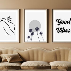 May include: Three framed art prints. The left print features a black line drawing of a person. The center print shows a gray sun and palm trees. The right print says "Good Vibes" in black script. The prints are above a beige sofa.