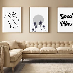 May include: Three framed art prints with gold frames hang above a beige sofa. The prints feature minimalist line art, a palm tree design with a gray sun, and the words "Good Vibes" in a bold, black font. The sofa has a tufted back and a neutral color.