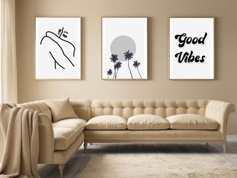 May include: Three framed art prints with gold frames hang above a beige sofa. The prints feature minimalist line art, a palm tree design with a gray sun, and the words "Good Vibes" in a bold, black font. The sofa has a tufted back and a neutral color.