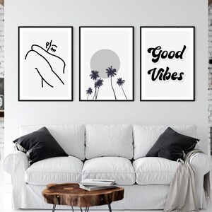 May include: Three framed wall art prints. The left print features a black line drawing of a person embracing themselves. The center print shows a gray sun and black palm trees. The right print says "Good Vibes" in black script. The prints are above a white couch.