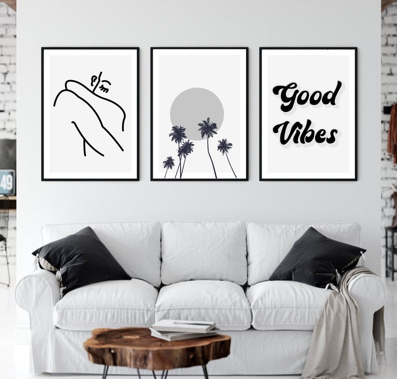 May include: Three framed wall art prints. The left print features a black line drawing of a person embracing themselves. The center print shows a gray sun and black palm trees. The right print says "Good Vibes" in black script. The prints are above a white couch.